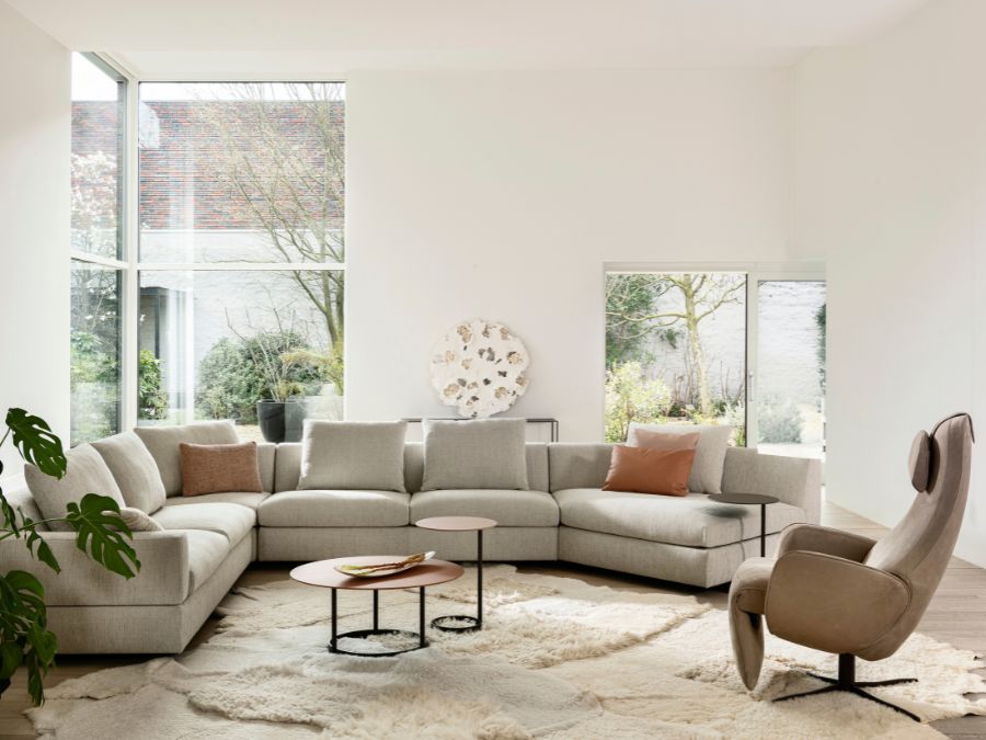 Lichtgrijze Jori Tigra Divanbase hoeksalon met roze en terracotta kussens, ronde salontafels en een taupe relaxfauteuil op een wollig tapijt Lichtgrijze Jori Tigra Divanbase hoeksalon met roze en terracotta kussens, ronde salontafels en een taupe relaxfauteuil op een wollig tapijtv