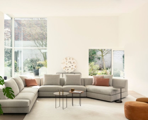 Ruime Jori Tigra Divanbase hoekzetel in lichtgrijze stof met twee kleine glazen en houten bijzettafels en cognackleurige poefs, in een woonkamer met grote ramen naar de tuin