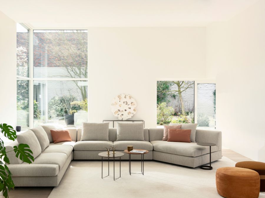 Ruime Jori Tigra Divanbase hoekzetel in lichtgrijze stof met twee kleine glazen en houten bijzettafels en cognackleurige poefs, in een woonkamer met grote ramen naar de tuin Ruime Jori Tigra Divanbase hoekzetel in lichtgrijze stof met twee kleine glazen en houten bijzettafels en cognackleurige poefs, in een woonkamer met grote ramen naar de tuin