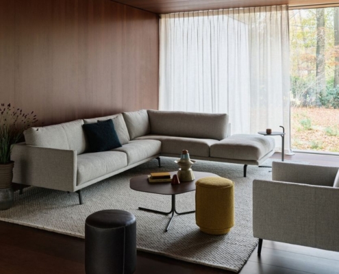 Leolux Bellice hoekbank in lichtgrijze stof met slanke metalen poten, met bijpassende fauteuil en poefs in een warme woonkamer met houten wanden