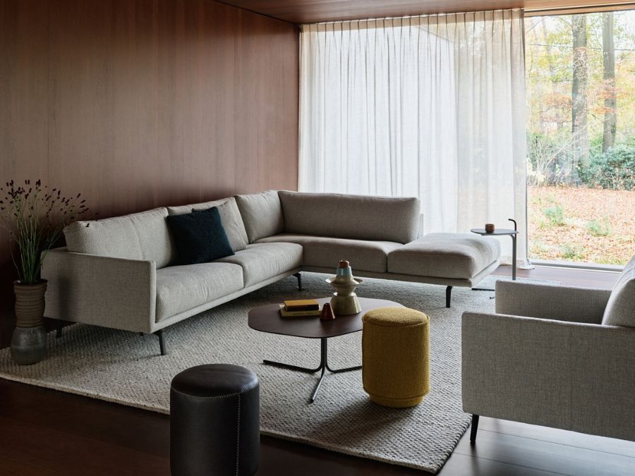 Leolux Bellice hoekbank in lichtgrijze stof met slanke metalen poten, met bijpassende fauteuil en poefs in een warme woonkamer met houten wanden