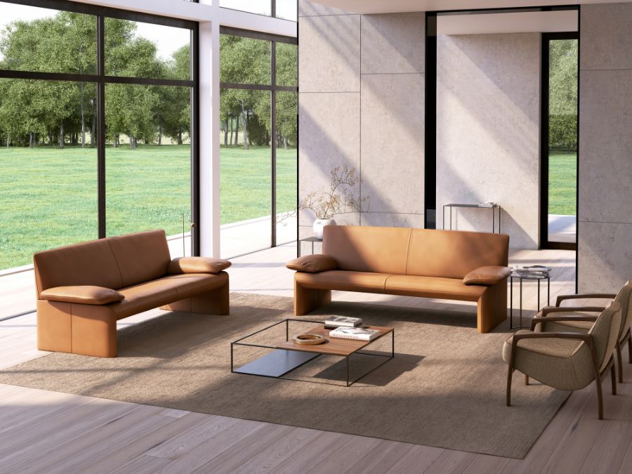 Twee cognac lederen Jori Linea zitbanken met open onderkant, schuin opgesteld rond een lage salontafel in een ruime woonkamer met glazen gevel Twee cognac lederen Jori Linea zitbanken met open onderkant, schuin opgesteld rond een lage salontafel in een ruime woonkamer met glazen gevel