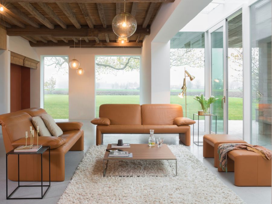 Twee Jori Linea zetels en bijpassende poefs in cognac leder rond een salontafel, in een lichte woonkamer met houten balken en zicht op het groen Twee Jori Linea zetels en bijpassende poefs in cognac leder rond een salontafel, in een lichte woonkamer met houten balken en zicht op het groen